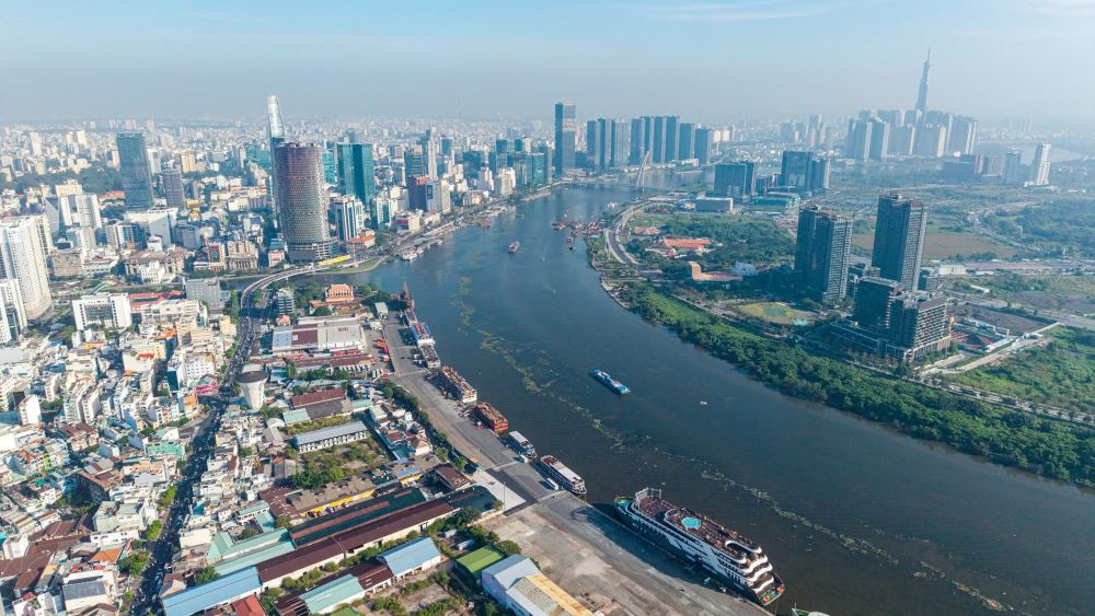 [Ảnh 1: Góc máy flycam bao quát từ trên cao bóc tách rõ rệt sự tương phản bề mặt đô thị, một bên là những mái tôn xỉn màu bám bụi thời gian của thương cảng Khánh Hội hiện hữu, một bên là nhịp độ phát triển đang nén chặt chờ được khơi thông.]